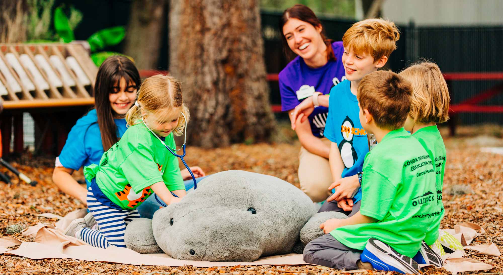 A group of young children learn from an instructor at ZooTampa Summer Camp how to perform a medical checkup on a stuffed manatee